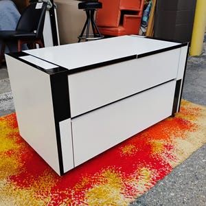 May include: White dresser with two drawers and black trim. The dresser is sitting on a colorful rug.