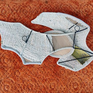 May include: An abstract textile art piece in light grey, with a textured surface and a complex, layered design. It's constructed from various materials and supported by a black metal frame. The artwork is set against an orange background.