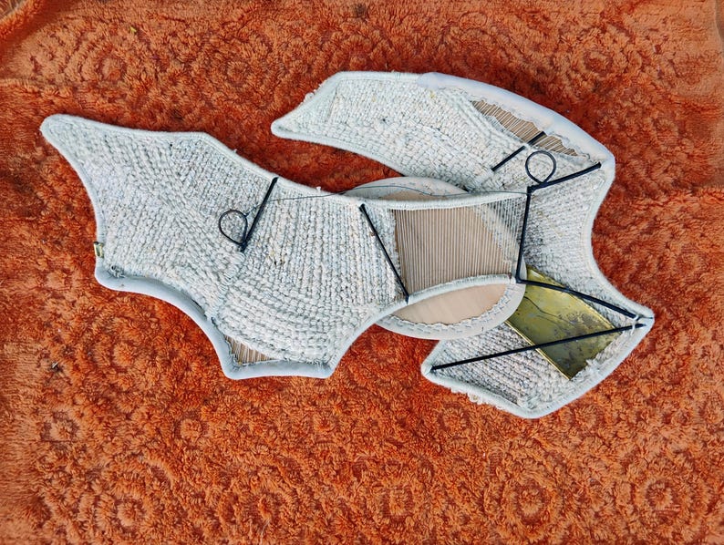 May include: An abstract textile art piece in light grey, with a textured surface and a complex, layered design. It's constructed from various materials and supported by a black metal frame. The artwork is set against an orange background.
