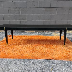May include: A rectangular coffee table with a glossy black finish, featuring four tapered black legs with gold-toned metal accents at the top and bottom. The table has a smooth, flat surface and a clean, linear design with visible gold hardware.