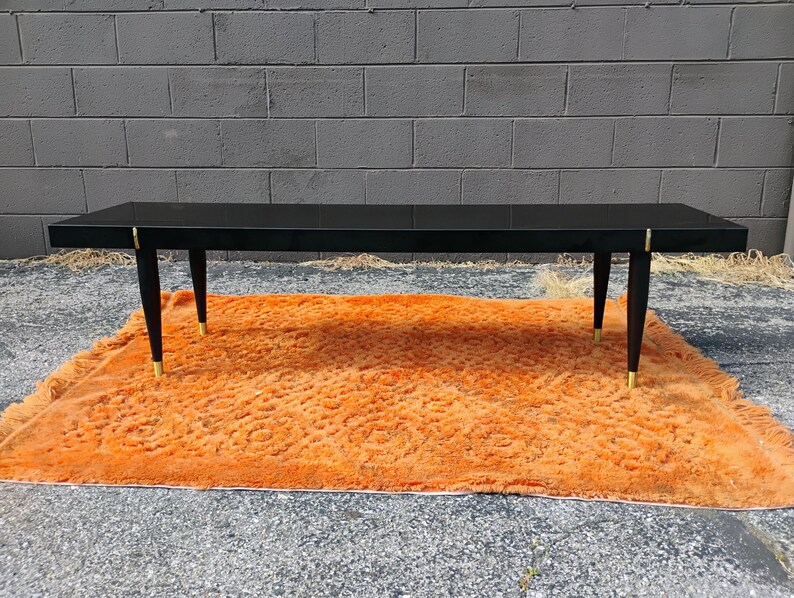 May include: A rectangular coffee table with a glossy black finish, featuring four tapered black legs with gold-toned metal accents at the top and bottom. The table has a smooth, flat surface and a clean, linear design with visible gold hardware.