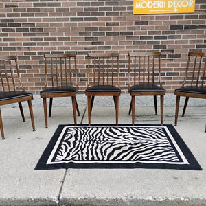 Puede incluir: Cinco sillas de madera con respaldos de varillas y asientos tapizados en negro dispuestas en fila, acompa&ntilde;adas de una alfombra rectangular con patr&oacute;n de rayas de cebra en blanco y negro y borde grueso negro, frente a una pared de ladrillo.