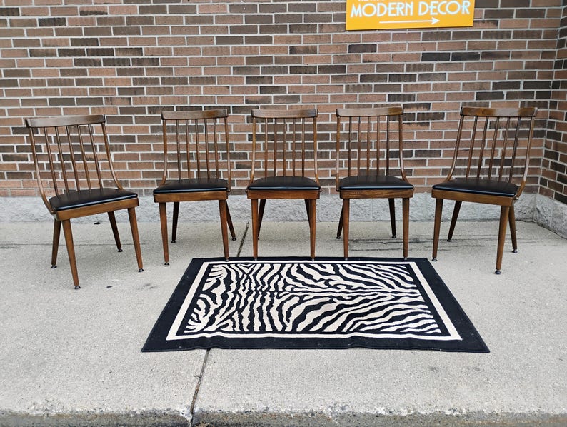 Puede incluir: Cinco sillas de madera con respaldos de varillas y asientos tapizados en negro dispuestas en fila, acompa&ntilde;adas de una alfombra rectangular con patr&oacute;n de rayas de cebra en blanco y negro y borde grueso negro, frente a una pared de ladrillo.