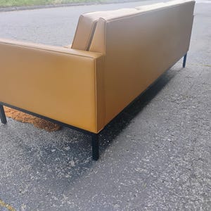 May include: A mustard yellow sofa with a smooth, solid-colored upholstery finish. The sofa features a rectangular frame with clean, straight lines and is supported by four thin, black metal legs positioned at each corner of the base structure.