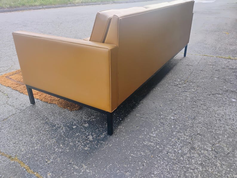 May include: A mustard yellow sofa with a smooth, solid-colored upholstery finish. The sofa features a rectangular frame with clean, straight lines and is supported by four thin, black metal legs positioned at each corner of the base structure.
