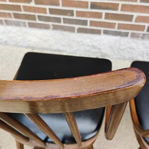 Puede incluir: Un par de sillas de madera con respaldos curvos en acabado marr&oacute;n medio y asientos tapizados en negro. Los respaldos est&aacute;n construidos con barrotes verticales y muestran una textura de veta de madera visible y desgaste.