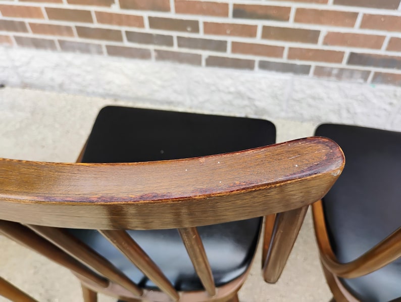 Puede incluir: Un par de sillas de madera con respaldos curvos en acabado marr&oacute;n medio y asientos tapizados en negro. Los respaldos est&aacute;n construidos con barrotes verticales y muestran una textura de veta de madera visible y desgaste.