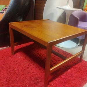 May include: A rectangular wooden coffee table with a light brown finish. The table has a simple design with a flat top and four legs. The table is sitting on a red shag rug.