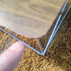 May include: A small table with a light brown wood-grain top and a thin, white metal wire frame base. The table features a square surface with rounded corners and a minimalist structure supported by four slender white metal legs.