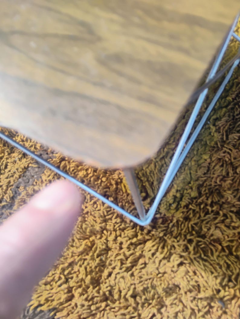 May include: A small table with a light brown wood-grain top and a thin, white metal wire frame base. The table features a square surface with rounded corners and a minimalist structure supported by four slender white metal legs.