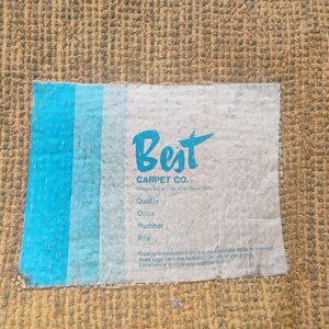 Puede incluir: Una alfombra texturizada de color tostado con una etiqueta rectangular blanca que presenta rayas verticales azules en el lado izquierdo. La etiqueta muestra el texto Best CARPET CO. junto con campos para Quality, Color, Number y Pile.