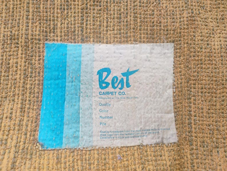 Puede incluir: Una alfombra texturizada de color tostado con una etiqueta rectangular blanca que presenta rayas verticales azules en el lado izquierdo. La etiqueta muestra el texto Best CARPET CO. junto con campos para Quality, Color, Number y Pile.