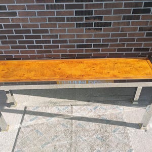K&ouml;nnte beinhalten: Ein rechteckiger Couchtisch mit polierter Holzplatte und Edelstahlrahmen. Die Holzplatte hat eine warme, goldbraune Farbe mit sichtbarem Holzmaserungsmuster. Der Tisch ist 152 cm lang und 51 cm breit.