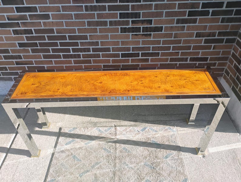 K&ouml;nnte beinhalten: Ein rechteckiger Couchtisch mit polierter Holzplatte und Edelstahlrahmen. Die Holzplatte hat eine warme, goldbraune Farbe mit sichtbarem Holzmaserungsmuster. Der Tisch ist 152 cm lang und 51 cm breit.