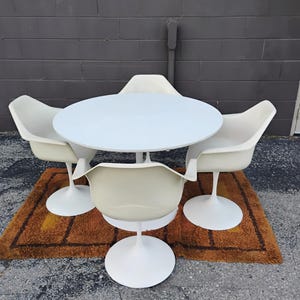 May include: A white circular dining table with a central pedestal base, accompanied by four matching off-white molded armchairs, each featuring a single white pedestal base and a curved, continuous seat and backrest design.