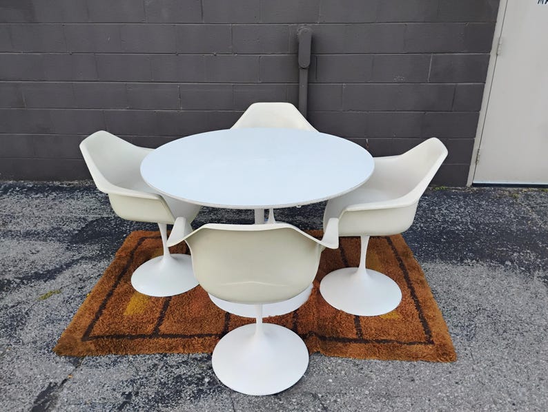 May include: A white circular dining table with a central pedestal base, accompanied by four matching off-white molded armchairs, each featuring a single white pedestal base and a curved, continuous seat and backrest design.