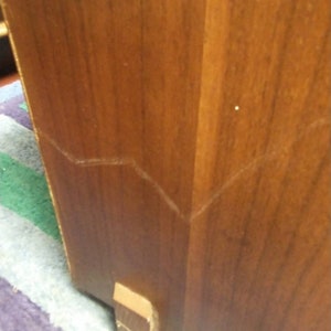 May include: Close-up of a wooden cabinet corner. The cabinet is a warm brown color with visible wood grain. A small, rectangular wooden foot is attached to the bottom. The cabinet has a visible seam.