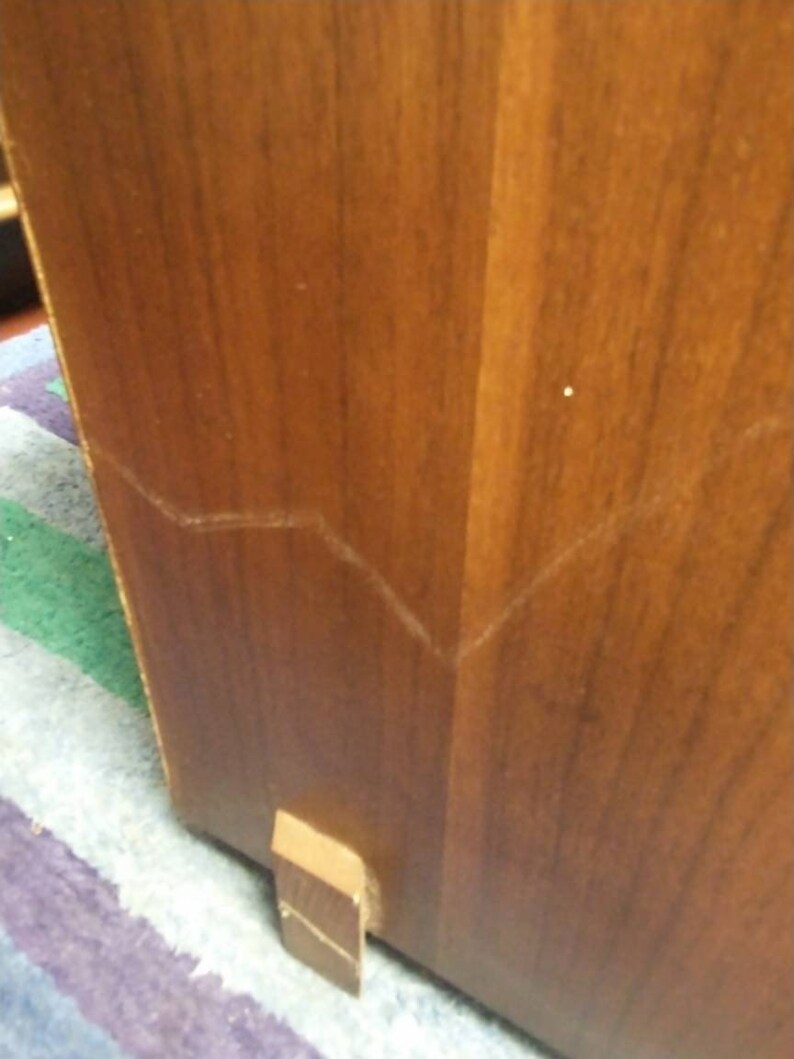 May include: Close-up of a wooden cabinet corner. The cabinet is a warm brown color with visible wood grain. A small, rectangular wooden foot is attached to the bottom. The cabinet has a visible seam.