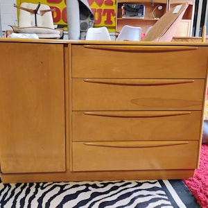 May include: A light wood dresser with four drawers and a cabinet door on the left side. The dresser has a curved top and rounded edges.