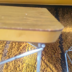 May include: A rectangular table with a light brown wood-grain top and a smooth yellow edge, supported by thin, silver-colored metal wire legs. The table surface shows minor wear along the rounded corner edge, and the metal frame is visible.