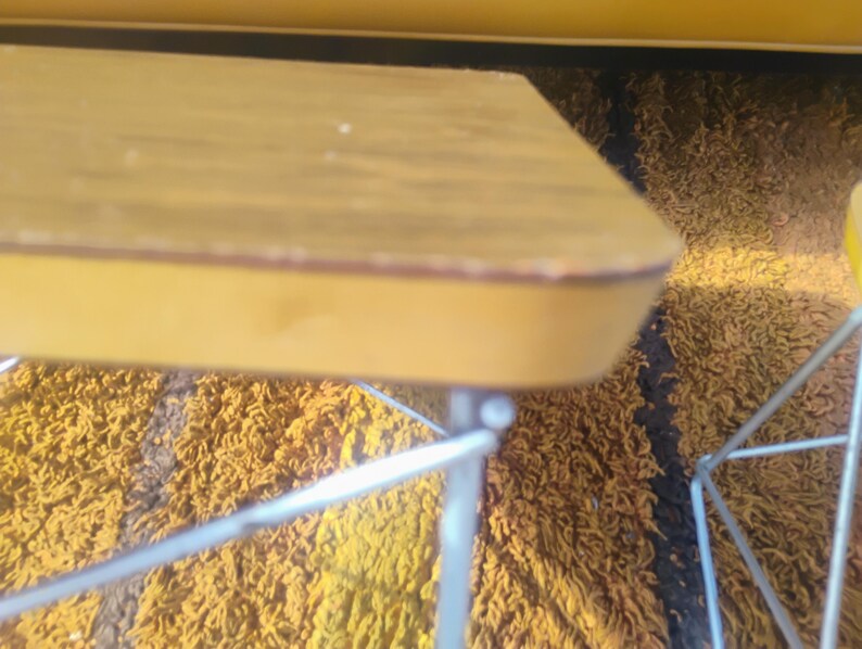 May include: A rectangular table with a light brown wood-grain top and a smooth yellow edge, supported by thin, silver-colored metal wire legs. The table surface shows minor wear along the rounded corner edge, and the metal frame is visible.