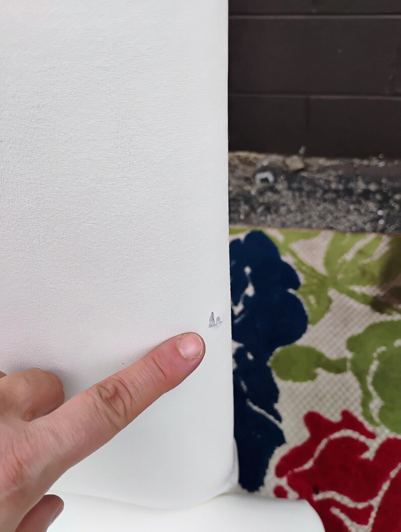 May include: A close-up of a white object with a textured surface. A finger is pointing at a small tear on the object. The background includes a floral patterned rug with red, blue, and green flowers.