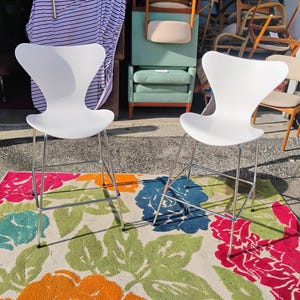 K&ouml;nnte beinhalten: Zwei wei&szlig;e, moderne Barhocker mit Chrombeinen stehen auf einem farbenfrohen Blummen-Teppich. Die Hocker haben ein markantes, geschwungenes R&uuml;cken- und Sitzdesign. Der Teppich zeigt gro&szlig;e Blumen in Rosa, Orange, Blau und Gr&uuml;n.