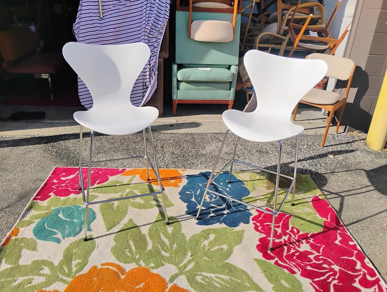 K&ouml;nnte beinhalten: Zwei wei&szlig;e, moderne Barhocker mit Chrombeinen stehen auf einem farbenfrohen Blummen-Teppich. Die Hocker haben ein markantes, geschwungenes R&uuml;cken- und Sitzdesign. Der Teppich zeigt gro&szlig;e Blumen in Rosa, Orange, Blau und Gr&uuml;n.