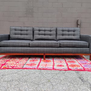 K&ouml;nnte beinhalten: Ein Dreisitzer-Sofa mit dunkelgrauem, strukturiertem Stoffbezug und einem dezenten Karomuster. Das Sofa verf&uuml;gt &uuml;ber getuftete R&uuml;ckenkissen, gerade Armlehnen und einen h&ouml;lzernen Unterbau mit vier Beinen, was f&uuml;r ein strukturiertes Erscheinungsbild sorgt.