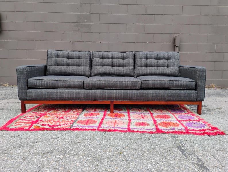 K&ouml;nnte beinhalten: Ein Dreisitzer-Sofa mit dunkelgrauem, strukturiertem Stoffbezug und einem dezenten Karomuster. Das Sofa verf&uuml;gt &uuml;ber getuftete R&uuml;ckenkissen, gerade Armlehnen und einen h&ouml;lzernen Unterbau mit vier Beinen, was f&uuml;r ein strukturiertes Erscheinungsbild sorgt.
