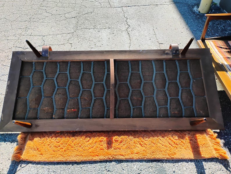 May include: A rectangular, mid-century modern speaker cover with a dark wood frame. The speaker cover has a brown mesh interior with a blue-gray honeycomb pattern. The cover is resting on an orange rug.