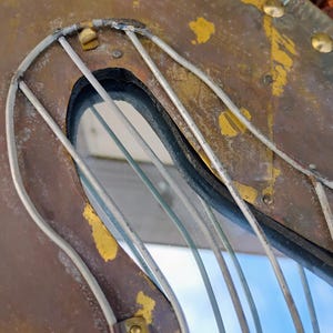 K&ouml;nnte beinhalten: Nahaufnahme eines Vintage-Spiegels in Form einer Harfe aus verwittertem Metall. Der Rahmen ist eine Mischung aus Braun- und Goldt&ouml;nen, mit sichtbaren Nieten und einer drahtartigen Struktur, die die Harfensaiten bildet. Der Spiegel reflektiert einen blauen Himmel.