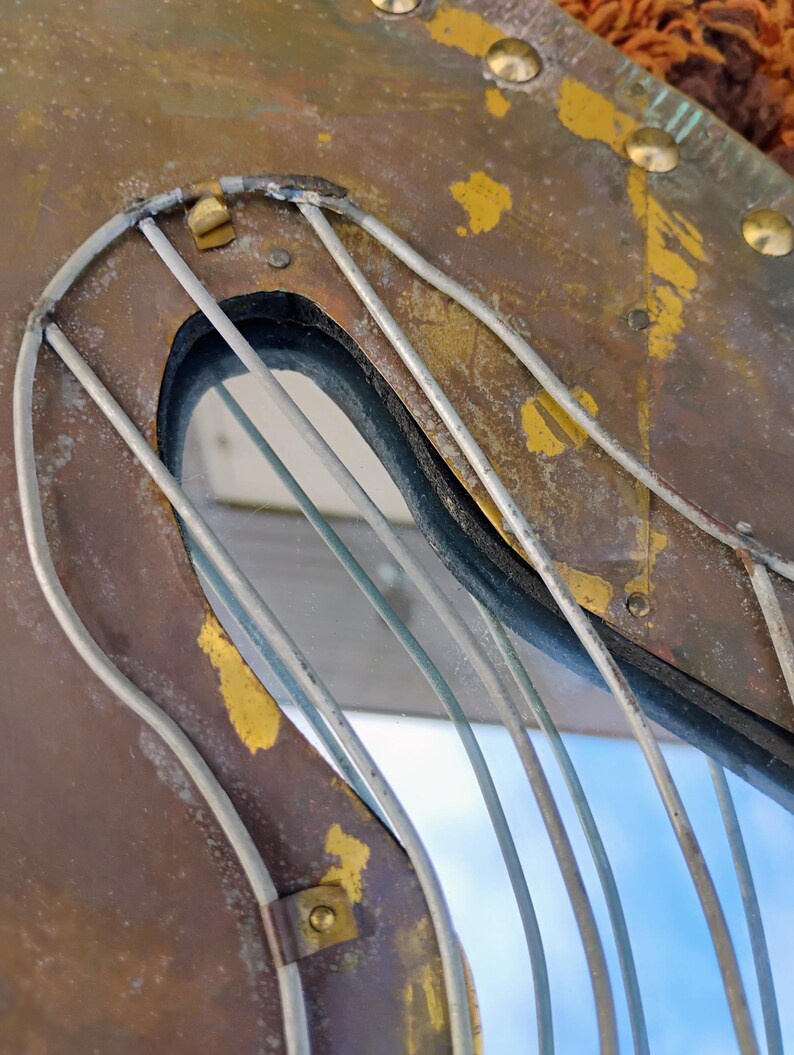 K&ouml;nnte beinhalten: Nahaufnahme eines Vintage-Spiegels in Form einer Harfe aus verwittertem Metall. Der Rahmen ist eine Mischung aus Braun- und Goldt&ouml;nen, mit sichtbaren Nieten und einer drahtartigen Struktur, die die Harfensaiten bildet. Der Spiegel reflektiert einen blauen Himmel.