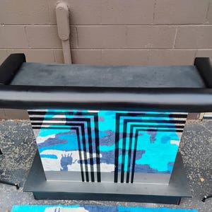 Pode incluir: Uma barra preta com uma parte superior acolchoada e uma base espelhada com um padrão geométrico em preto e azul. A barra está sentada em um tapete com um padrão semelhante.
