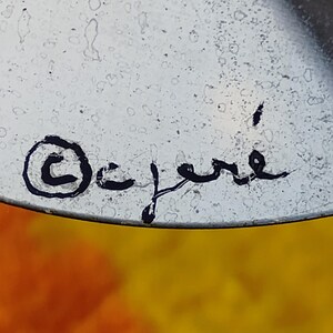 May include: A metallic surface featuring a black handwritten signature that reads C. Jere with a copyright symbol enclosed in a circle to the left. The surface has a slightly speckled texture and a reflective quality under the light.