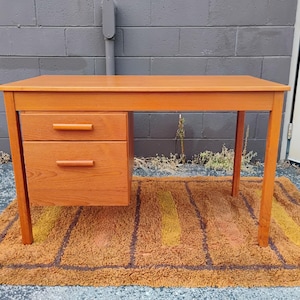 Op de afbeelding: Een rechthoekig houten bureau met een licht oranje-bruine afwerking, voorzien van een vlak bovenblad en een blok aan de linkerzijde met twee gestapelde lades, elk uitgerust met een horizontale handgreep in dezelfde kleur als het hout.