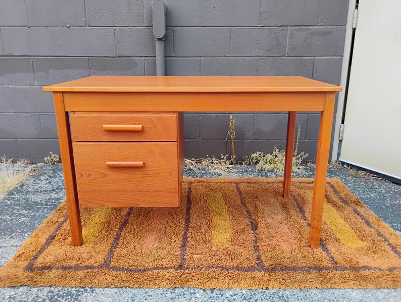 Op de afbeelding: Een rechthoekig houten bureau met een licht oranje-bruine afwerking, voorzien van een vlak bovenblad en een blok aan de linkerzijde met twee gestapelde lades, elk uitgerust met een horizontale handgreep in dezelfde kleur als het hout.