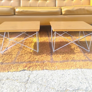 May include: Two rectangular side tables with light brown wooden tops and silver-colored metal wire bases featuring a crisscross support structure. The tables have smooth, flat surfaces and are positioned side by side on a textured surface.