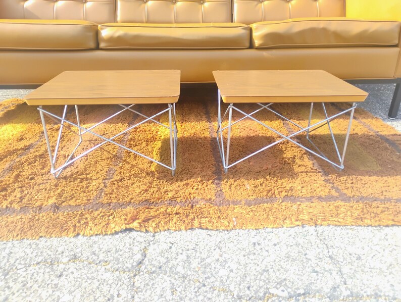 May include: Two rectangular side tables with light brown wooden tops and silver-colored metal wire bases featuring a crisscross support structure. The tables have smooth, flat surfaces and are positioned side by side on a textured surface.