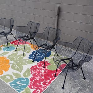 May include: Four black metal chairs with a wire mesh design are arranged on a white rug with a colorful floral pattern.