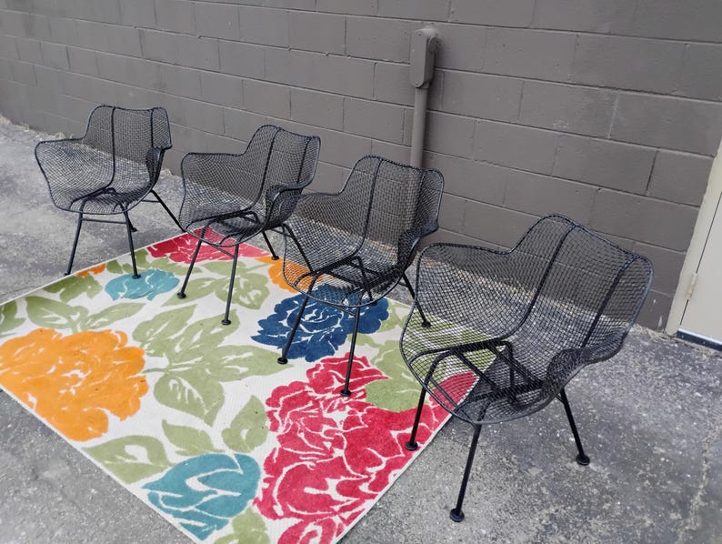 May include: Four black metal chairs with a wire mesh design are arranged on a white rug with a colorful floral pattern.