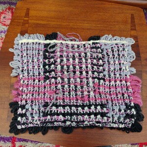 May include: A handwoven textile in shades of pink, black, gray, and white. The rectangular piece features a textured weave with fringed edges. The textile is displayed on a wooden surface, showcasing its intricate design and craftsmanship.