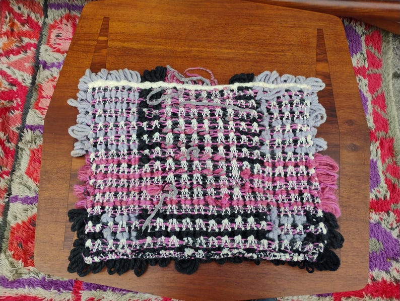 May include: A handwoven textile in shades of pink, black, gray, and white. The rectangular piece features a textured weave with fringed edges. The textile is displayed on a wooden surface, showcasing its intricate design and craftsmanship.