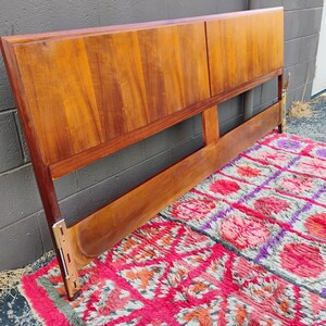 May include: A rectangular wooden headboard featuring a warm, medium-brown wood grain finish. The headboard consists of two large, smooth upper panels and a single horizontal lower support rail, with visible metal mounting brackets at the base.