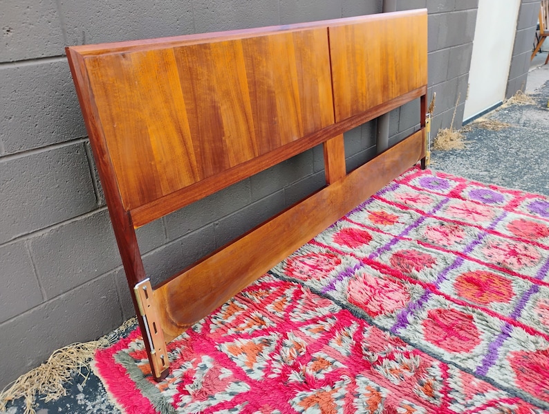 May include: A rectangular wooden headboard featuring a warm, medium-brown wood grain finish. The headboard consists of two large, smooth upper panels and a single horizontal lower support rail, with visible metal mounting brackets at the base.