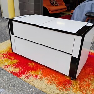 May include: A white and black two-drawer cabinet with a geometric design. The cabinet has a glossy finish and is sitting on a colorful rug.