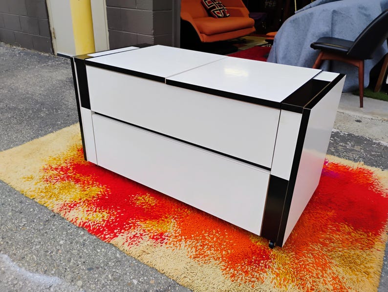 May include: A white and black two-drawer cabinet with a geometric design. The cabinet has a glossy finish and is sitting on a colorful rug.