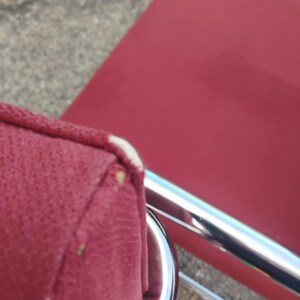 May include: A red upholstered chair featuring a chrome metal frame. The seat cushion has a visible white piping detail along the corner edge, and the fabric texture is clearly defined against the reflective metallic structure of the chair base.