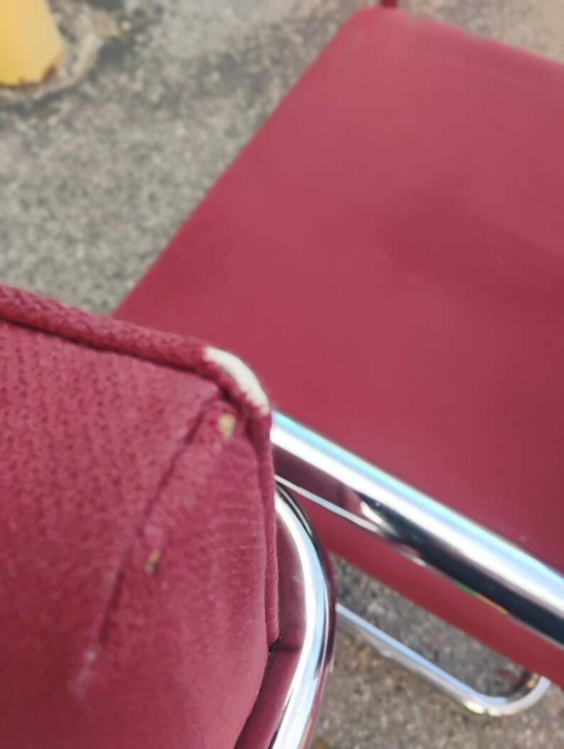 May include: A red upholstered chair featuring a chrome metal frame. The seat cushion has a visible white piping detail along the corner edge, and the fabric texture is clearly defined against the reflective metallic structure of the chair base.