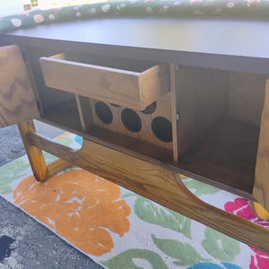 May include: A wooden cabinet with open doors, revealing a drawer and storage space. The cabinet has a dark brown top and light brown wooden doors and frame. The interior includes circular cutouts, likely for bottles. A colorful floral rug is visible below.
